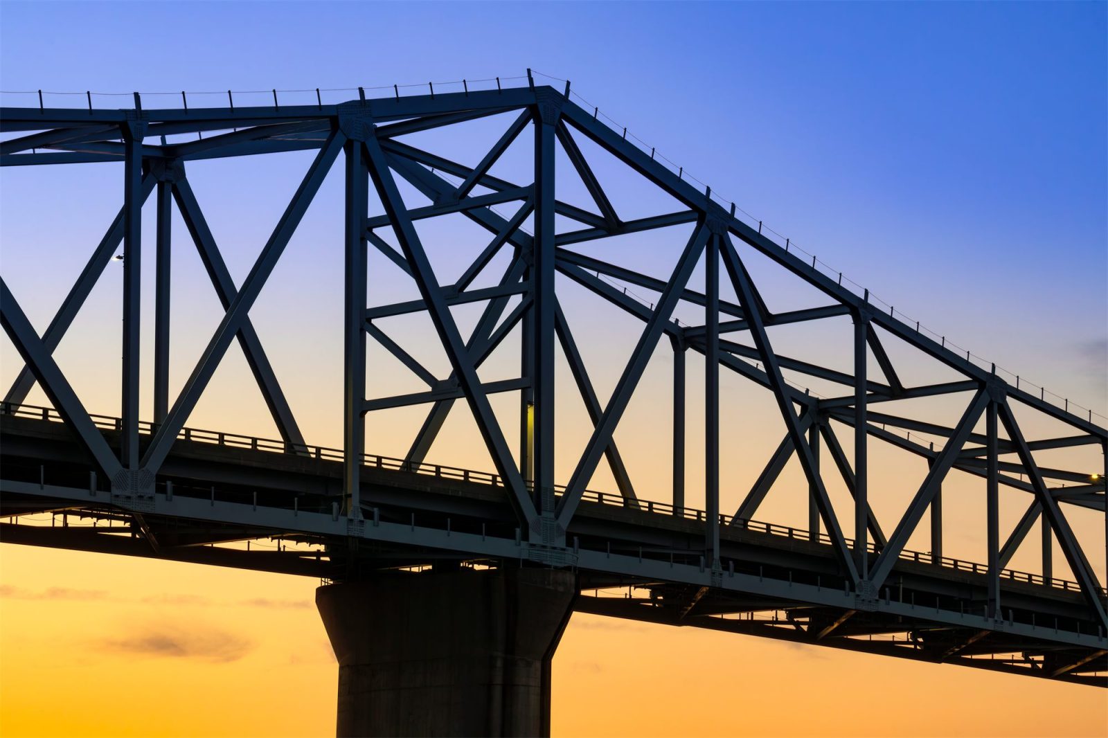 The steel trusses of the Milton–Madison Bridge silhouetted against an amber sunset over the Ohio River.